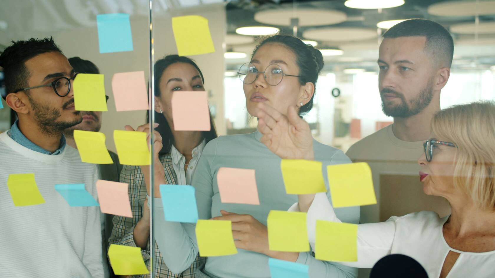 Tiimi seisoo lasiseinän takana, johon on kiinnitetty eri värisiä post it -lappuja.